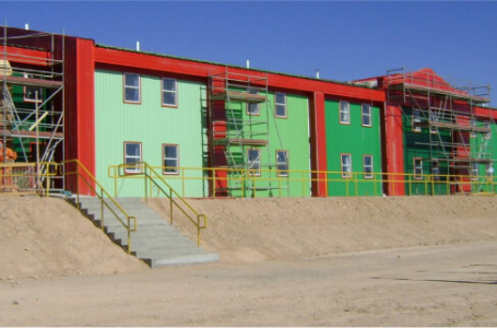 Edificio de viviendas modulares con un diseño colorido, que muestra una combinación de paredes verdes y rojas. Se aprecian escaleras y un entorno árido, ideal para una construcción rápida y eficiente. Esta imagen representa una opción de casa prefabricada versátil y funcional. Tecno Fast Home.