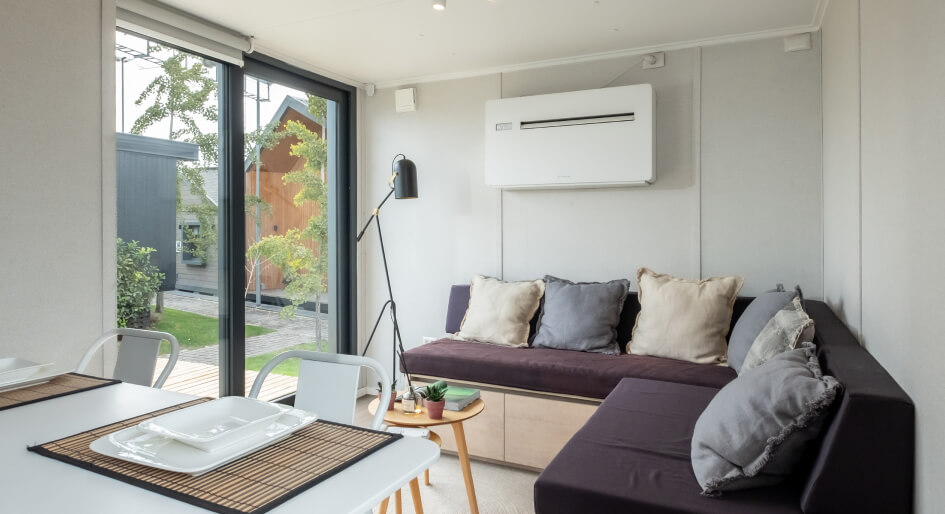 Un acogedor espacio interior de una vivienda modular, con un sof&aacute; c&oacute;modo y grandes ventanales que permiten la entrada de luz natural. Este dise&ntilde;o funcional es ideal para una casa prefabricada o una segunda vivienda. Tecno Fast Home.