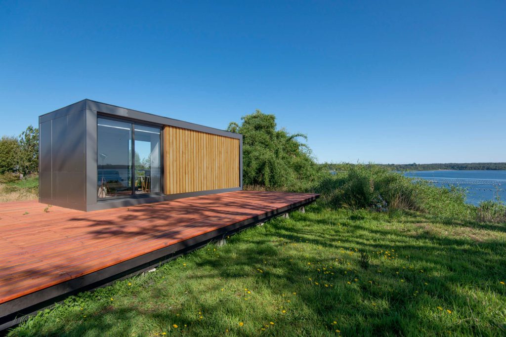 Una moderna vivienda modular con amplios ventanales, ubicada en un entorno natural, ideal como casa de vacaciones o segunda vivienda. Su diseño contemporáneo se integra perfectamente con el paisaje. Tecno Fast Home. --- Casa prefabricada con terraza de madera, rodeada de vegetación, perfecta para disfrutar de la naturaleza. Esta opción de vivienda modular ofrece comodidad y estilo. Tecno Fast Home. --- Espaciosa casa modular con exterior en madera, ideal para quienes buscan una segunda vivienda cerca del agua. Su estructura eficiente y estética moderna son características destacadas. Tecno Fast Home.