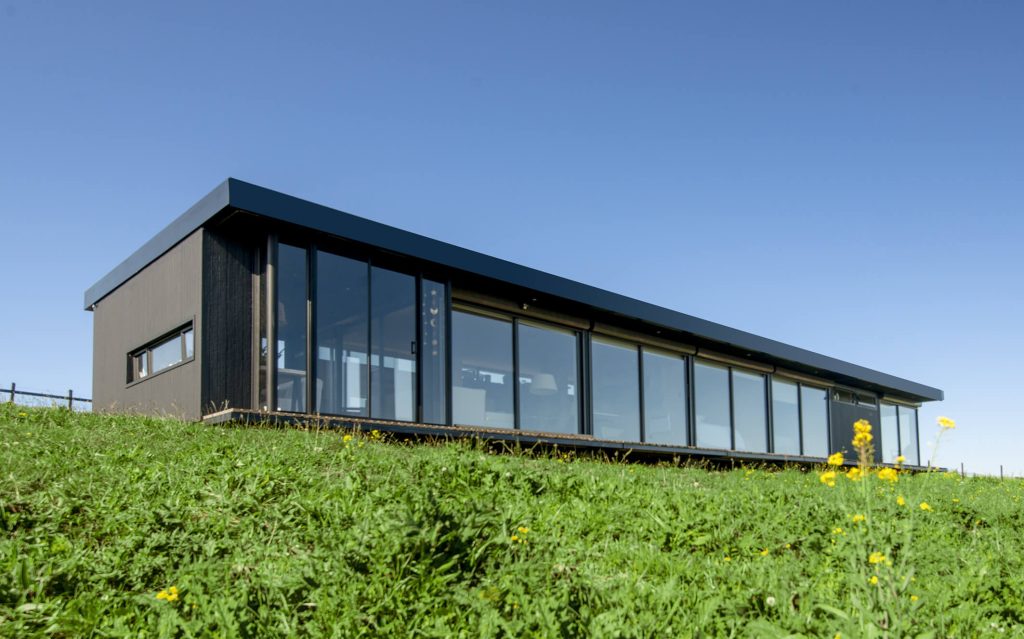 Casa modular moderna con grandes ventanales y un diseño elegante, ubicada en un entorno natural. Ideal como vivienda principal o segunda vivienda. Presenta un estilo contemporáneo que resalta la funcionalidad y la estética. Marca: Tecno Fast Home.