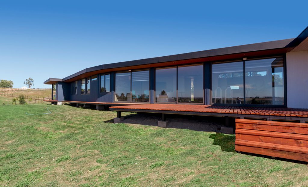 Casa modular moderna con amplias ventanas y una terraza de madera, ubicada en un entorno natural. Ideal como vivienda principal o segunda residencia. Diseño eficiente y contemporáneo. Tecno Fast Home.