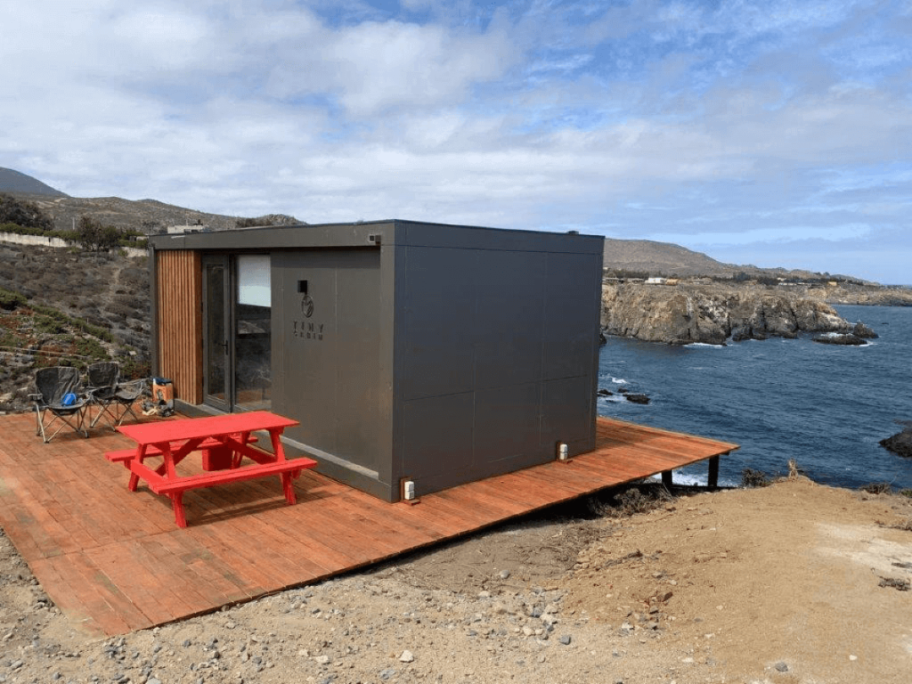 Tiny Cabin modular situada junto al mar, con un diseño contemporáneo y una terraza de madera, ideal como segunda vivienda. Esta vivienda prefabricada combina funcionalidad y estética, perfecta para disfrutar de la naturaleza. Marca Tecno Fast Home.