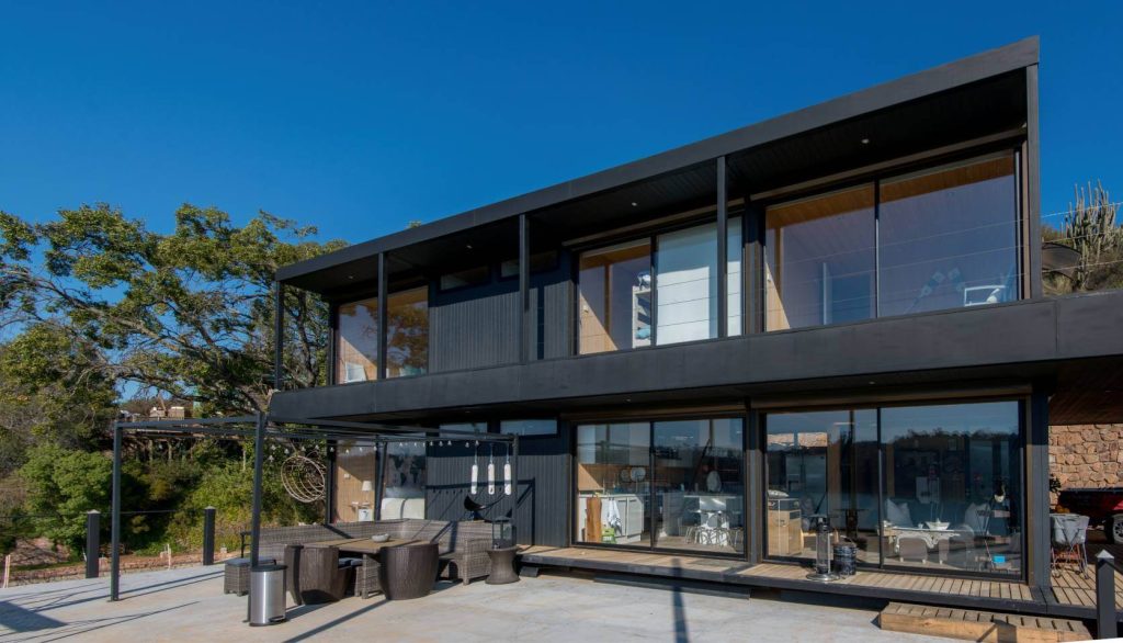 Casa modular de diseño contemporáneo con amplias ventanas y balcón, ideal para una segunda vivienda. Espacio exterior acogedor con muebles, perfecto para disfrutar al aire libre. Construcción de vivienda prefabricada que combina estética y funcionalidad. Tecno Fast Home.
