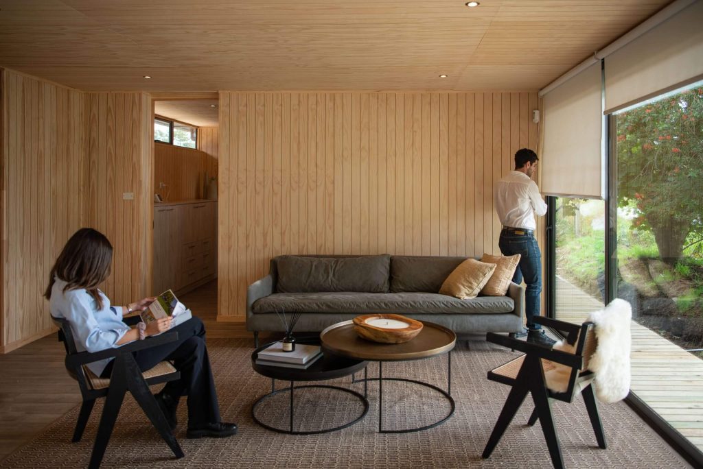 Un acogedor espacio interior de una vivienda modular, donde se observa un salón elegante con un sofá moderno y una mesa de centro. Una persona está sentada leyendo, mientras otra mira por la ventana, disfrutando de la vista. Este diseño representa la comodidad y funcionalidad de las casas prefabricadas. Tecno Fast Home.