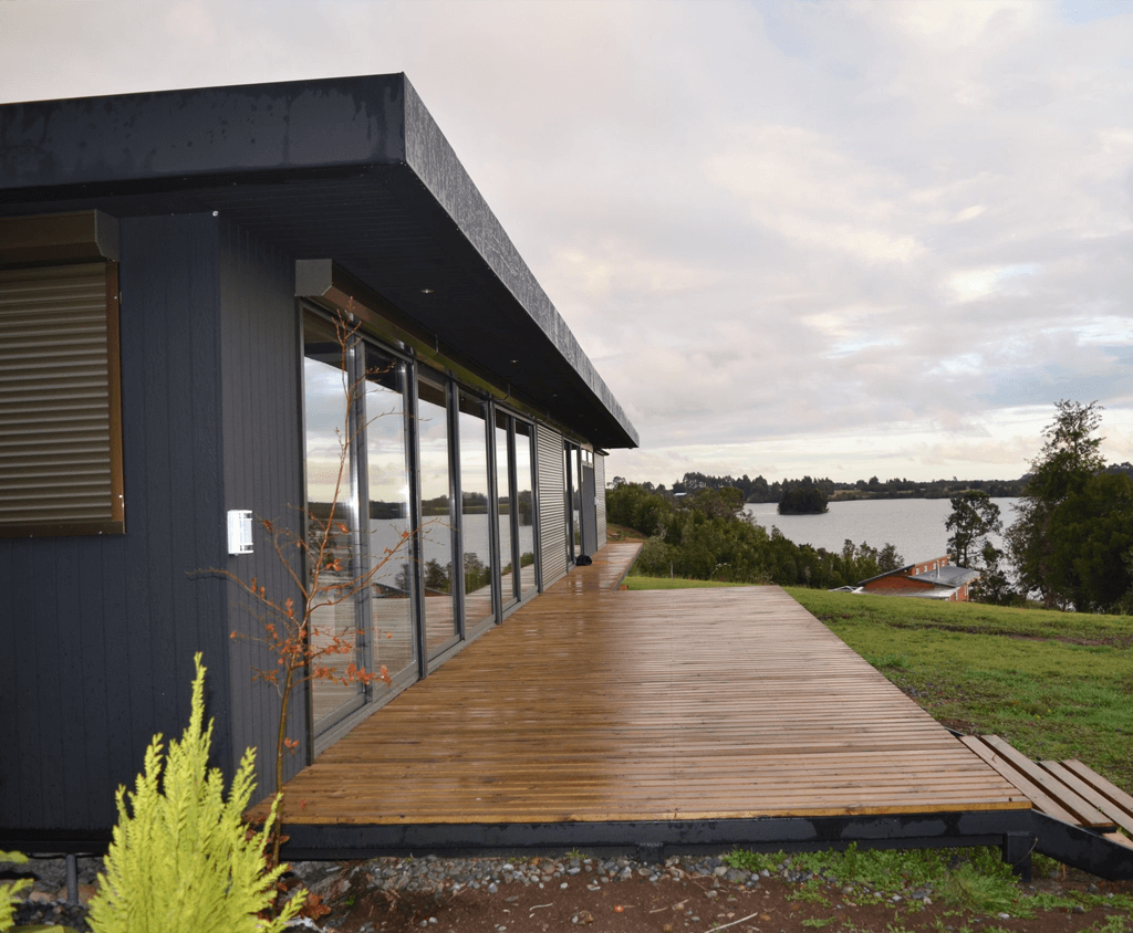 Casa modular moderna con amplias ventanas y terraza de madera, ubicada en un entorno natural junto al agua, ideal como segunda vivienda. Diseño elegante y funcional que destaca por su construcción eficiente. Tecno Fast Home.