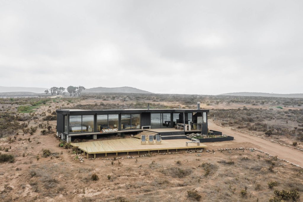 Casa modular contemporánea situada en un entorno desértico, con amplios ventanales que ofrecen vistas panorámicas y una terraza de madera. Ideal como vivienda modular o segunda vivienda. Diseño moderno y funcional de Tecno Fast Home.