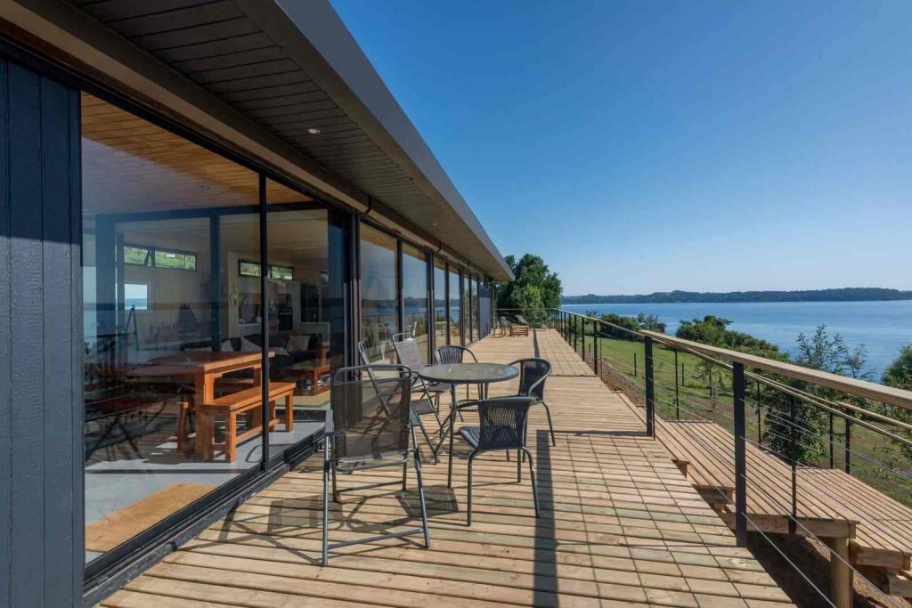 Amplia terraza de una vivienda modular con vistas al lago, diseñada para disfrutar del aire libre. La estructura presenta grandes ventanales que permiten la entrada de luz natural, ideal para una casa prefabricada. Espacio perfecto para una segunda vivienda o un refugio de vacaciones. Marca Tecno Fast Home.