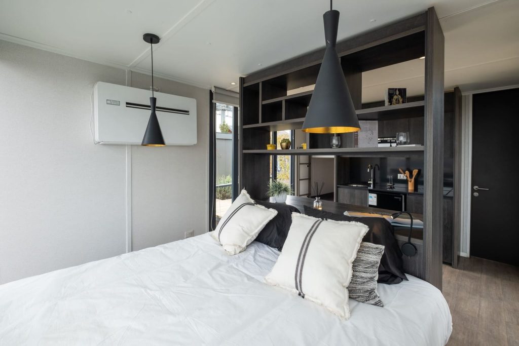 Habitación moderna y acogedora con cama y un diseño funcional, que incluye una pequeña cocina y área de trabajo, ideal para una vivienda modular o casa prefabricada. La iluminación y los detalles decorativos crean un ambiente cálido, perfecto para una segunda vivienda. Tecno Fast Home.