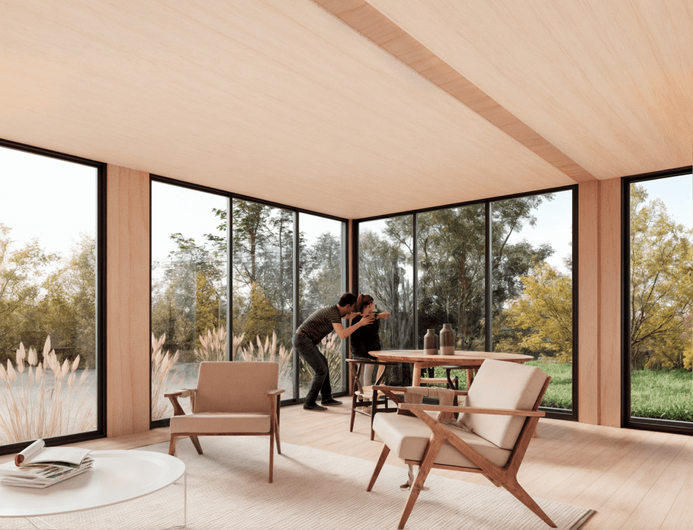 Interior de una casa modular con grandes ventanales que permiten la entrada de luz natural, mostrando un ambiente acogedor y moderno. Dos personas interactúan en un espacio que combina comodidad y funcionalidad, ideal para una vivienda prefabricada. Este diseño se destaca por su conexión con la naturaleza circundante. Tecno Fast Home.