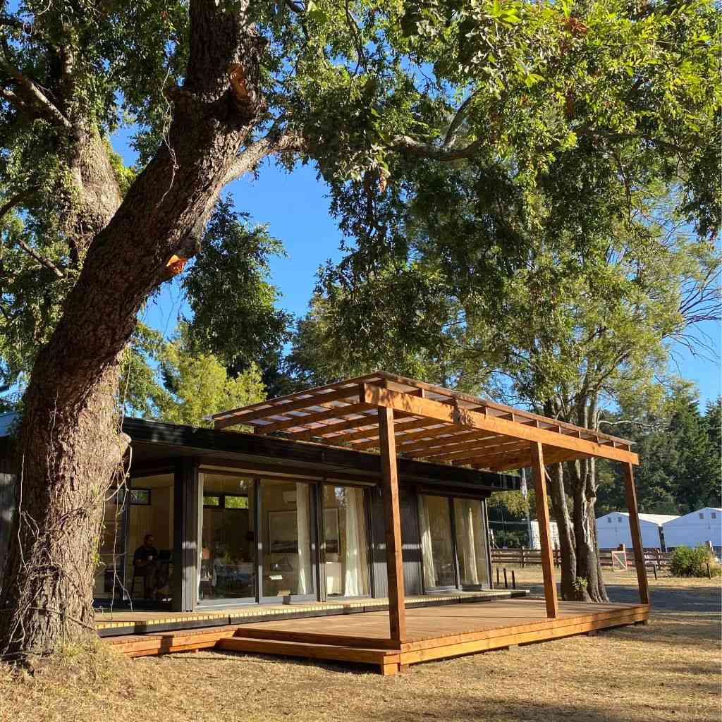 Casa modular rodeada de naturaleza, con una amplia terraza de madera que se extiende hacia un entorno verde. Esta vivienda prefabricada destaca por su diseño moderno y funcional, ideal para una segunda vivienda o como refugio. La marca Tecno Fast Home ofrece soluciones de vivienda innovadoras y sostenibles.