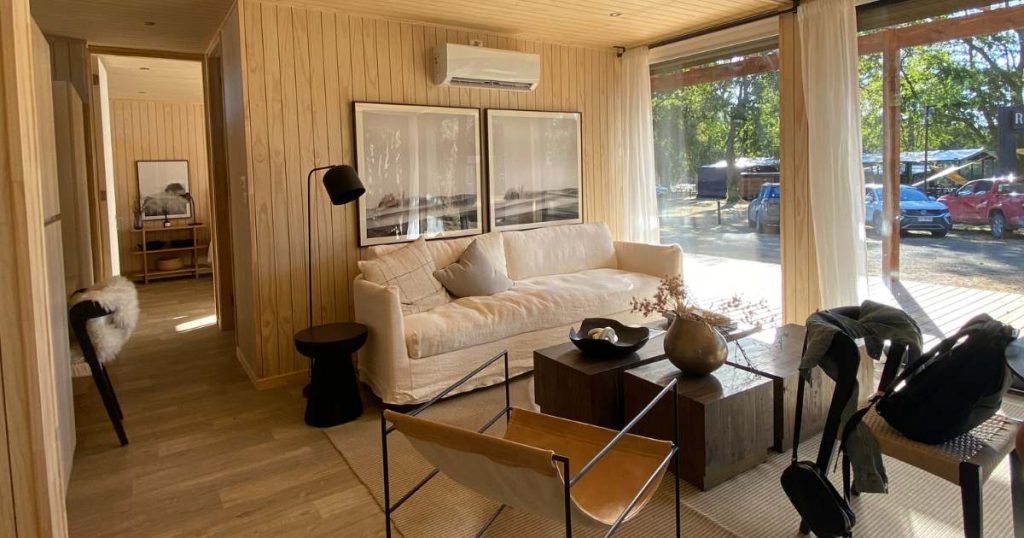 Interior de una vivienda modular, con un diseño contemporáneo y acogedor que incluye un sofá confortable y grandes ventanales que permiten la entrada de luz natural. La decoración es elegante y minimalista, ideal para una casa prefabricada. Este espacio es perfecto como segunda vivienda para disfrutar de la tranquilidad. Tecno Fast Home.