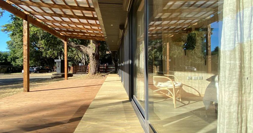 Una amplia terraza de madera que se extiende frente a una casa moderna, creando un espacio acogedor ideal para disfrutar de la naturaleza. La estructura cuenta con grandes ventanales que iluminan el interior de la vivienda. Este diseño de casa modular es perfecto como segunda vivienda o para escapadas. Marca: Tecno Fast Home.
Un área exteriores con un porche techado que conecta con una vivienda prefabricada, rodeada de árboles y vegetación. La casa destaca por su construcción eficiente y armon