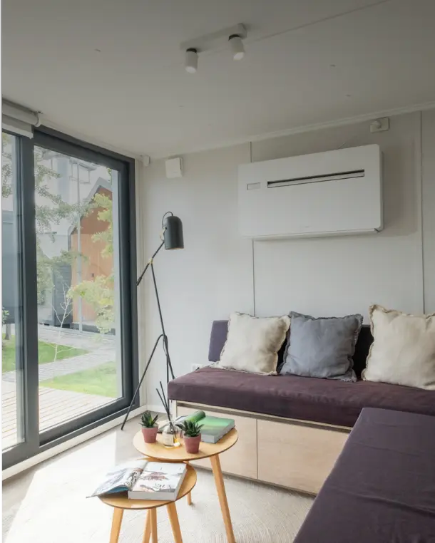Espacio interior acogedor de una vivienda modular, con un sofá cómodo y grandes ventanales que permiten la entrada de luz natural. Elementos decorativos como plantas y revistas en una mesa de centro complementan el ambiente. Ideal para disfrutar de una segunda vivienda. Tecno Fast Home.
Ambiente moderno y funcional de una casa prefabricada, donde se destaca un sofá con cojines y un sistema de aire acondicionado en la pared. Las amplias ventanas ofrecen vistas al exterior, creando un entorno luminos